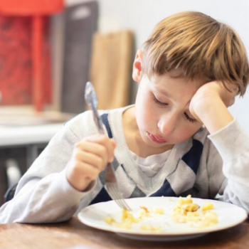 Child sitting at a kitchen table with a plate of food, holding a fork and resting head on one hand, appearing reluctant to eat.