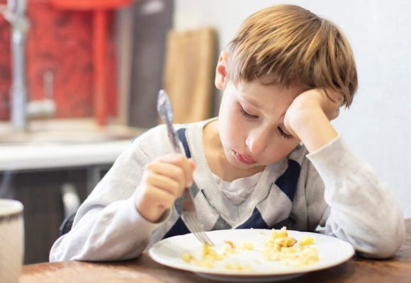 Child sitting at a kitchen table with a plate of food, holding a fork and resting head on one hand, appearing reluctant to eat.