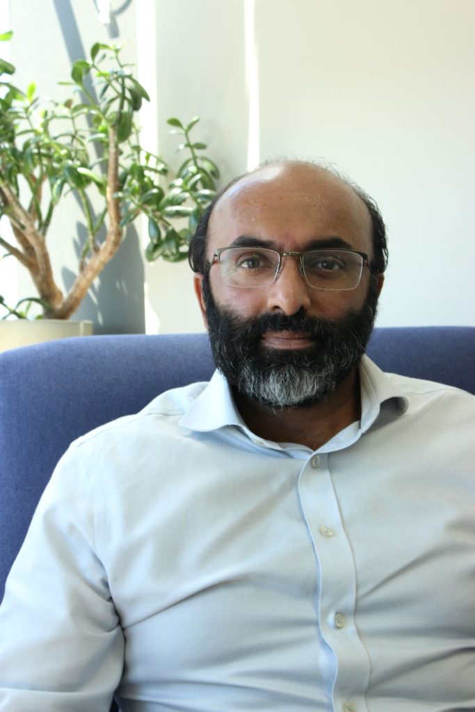 Dr Shyam Gangadharan, physician in General Medicine, seated indoors on a blue chair with a plant in the background.